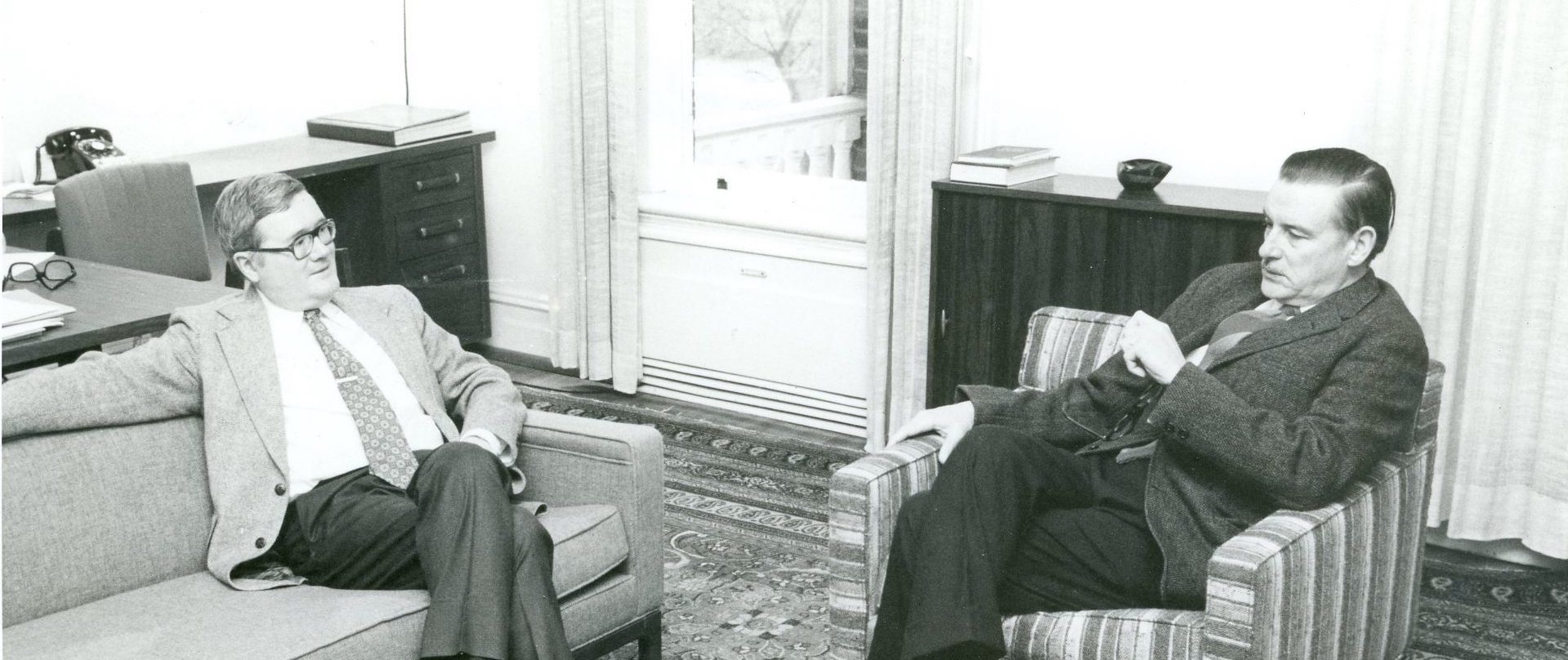 Black and white photograph. Gordon Tullock sits on a couch in an office listening. To his left, James Buchanan sits on a chair, speaking.